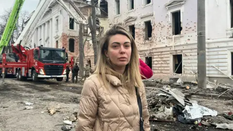 A woman named Nataliia looks straight into the camera with the remnants of her car pictured behind her. Fire engines are also stationed behind her, as well as damaged buildings.