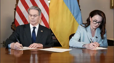 US Department of the Treasury US Treasury Secretary Scott Bessant wearing a suit and sitting in front of a US flag signs paper with a pen alongside Yulia Svyrydenko, Ukraine's First Deputy Prime Minister