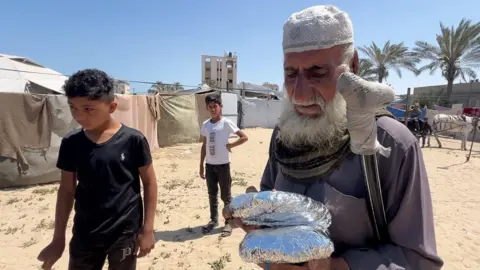 An elderly man holds two food parcels of food from an Anera community kitchen in the al-Mawasi area of southern Gaza